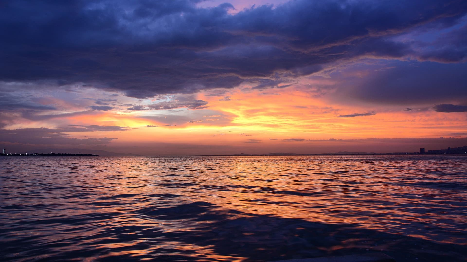 The sun on the horizon under a plum sky, over dark, calm water.