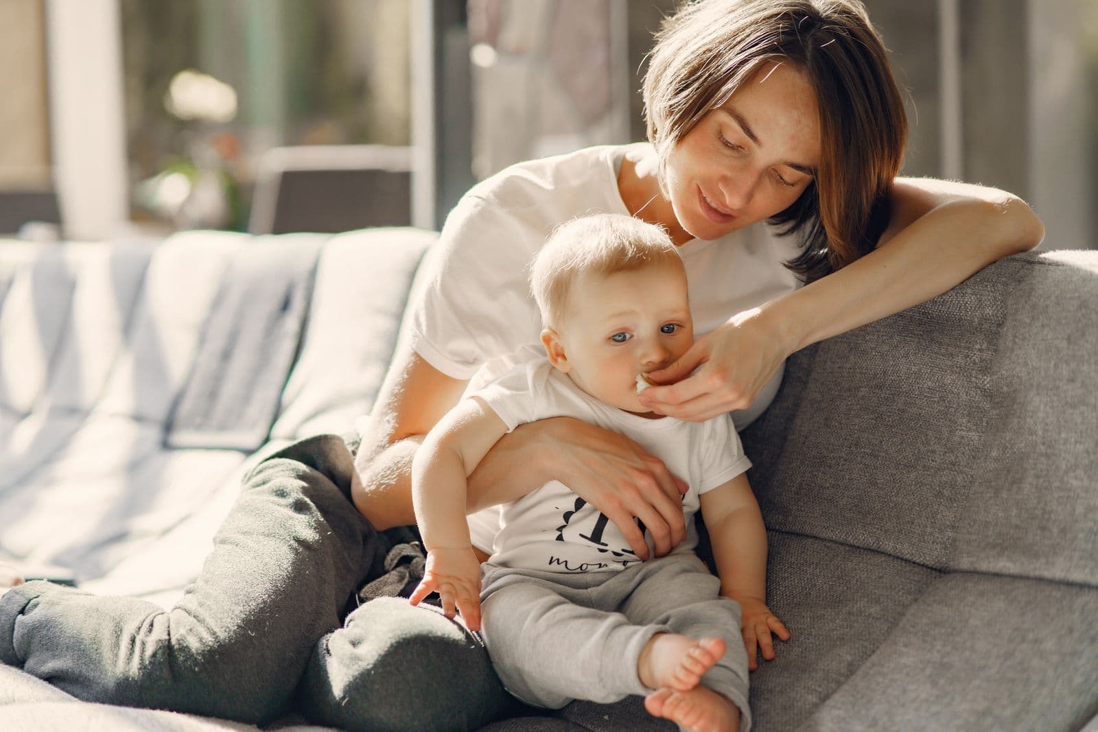 A mother holds her baby on a sofa in a sunlit living room.