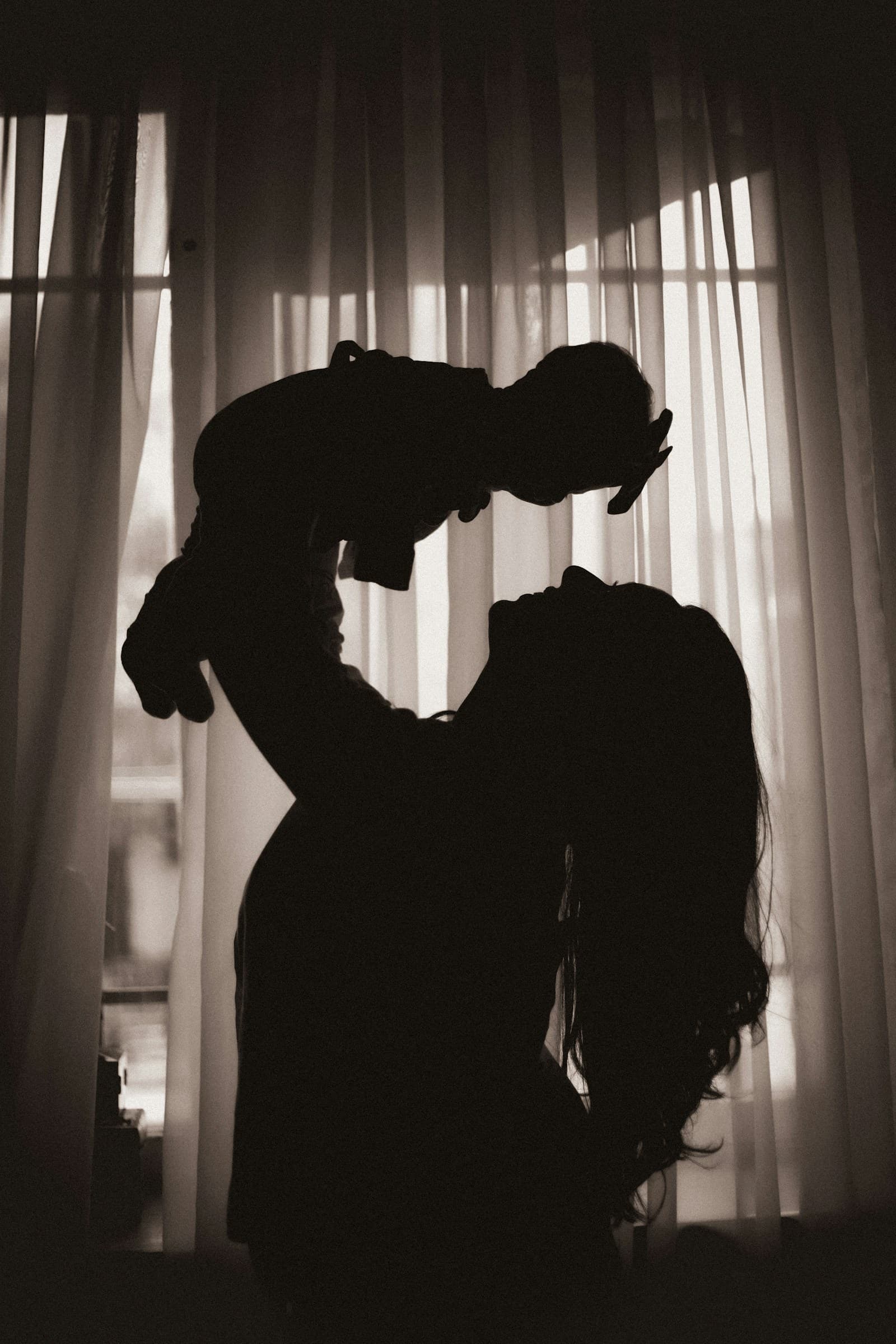 A woman lifts her baby in front of a bright window, in silhouette.